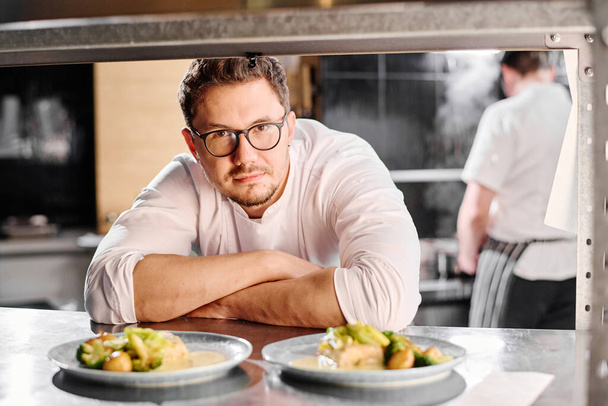 Portret młodego profesjonalnego kucharza w okularach patrzącego na kamerę za ladą z gotowymi daniami - Zdjęcie, obraz