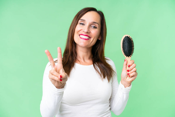 Kobieta w średnim wieku z grzebieniem włosów na odizolowanym tle klucza chroma uśmiechając się i pokazując znak zwycięstwa - Zdjęcie, obraz
