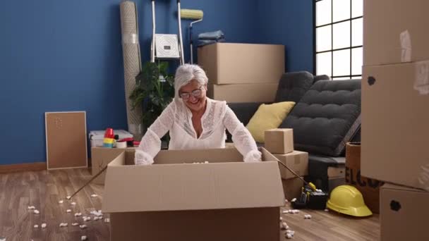 Mulher de cabelos grisalhos de meia idade sorrindo pacote unboxing confiante jogando com espuma de poliestireno em nova casa - Filmagem, Vídeo