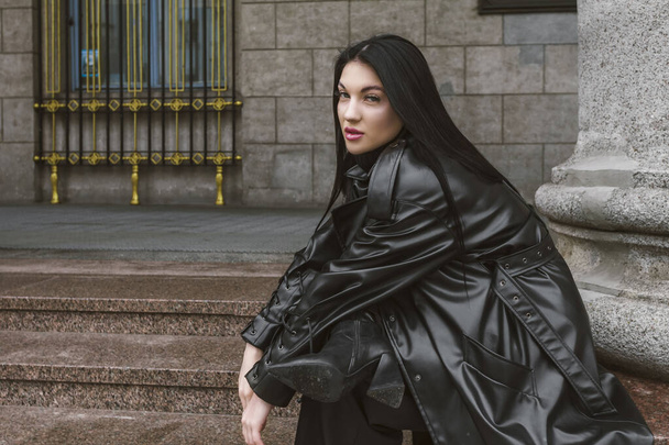 Straatfotografie. Een mooi stijlvol meisje gekleed in alle zwart, het dragen van een lange leren jas of jas, poseert tegen van grote gebouw kolommen. - Foto, afbeelding