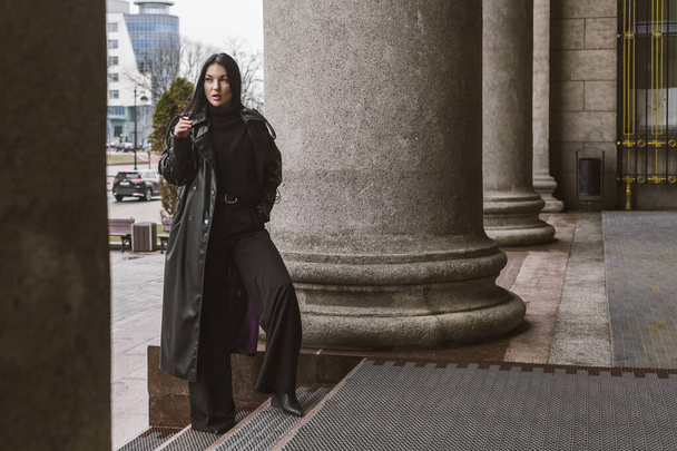 Straßenfotografie. Ein schönes, stylisches Mädchen ganz in schwarz gekleidet, trägt einen langen Ledermantel oder eine Jacke und posiert vor großen Bausäulen. - Foto, Bild