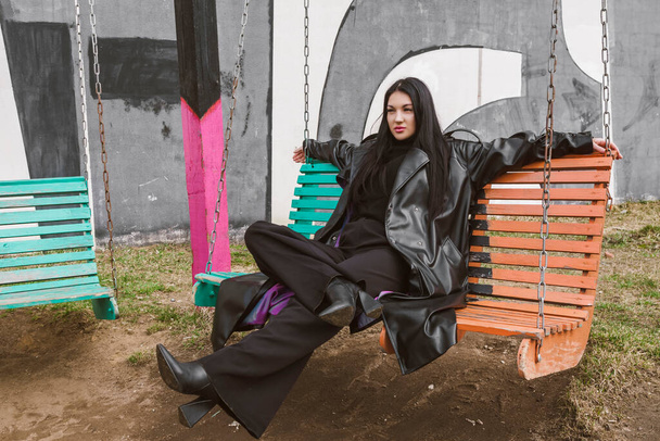 Fotografía callejera. Una hermosa chica vestida de negro, con un abrigo de cuero largo o chaqueta, se sienta en un banco oscilante frente a una pared cubierta de graffiti. - Foto, imagen