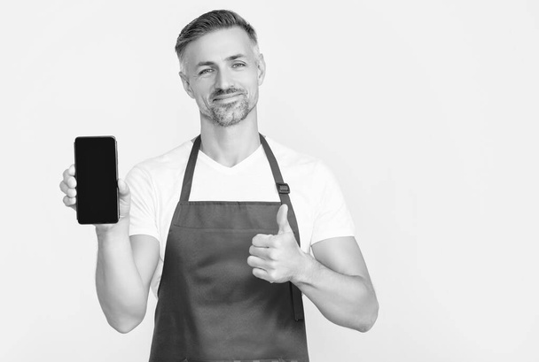 cheerful mature man in apron presenting smartphone with copy space. thumb up. - Фото, изображение