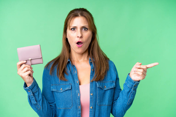 Middle age caucasian woman holding wallet over isolated background surprised and pointing side - Photo, Image