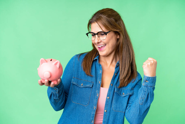 Middle age caucasian woman holding a piggybank over isolated background celebrating a victory - Фото, зображення