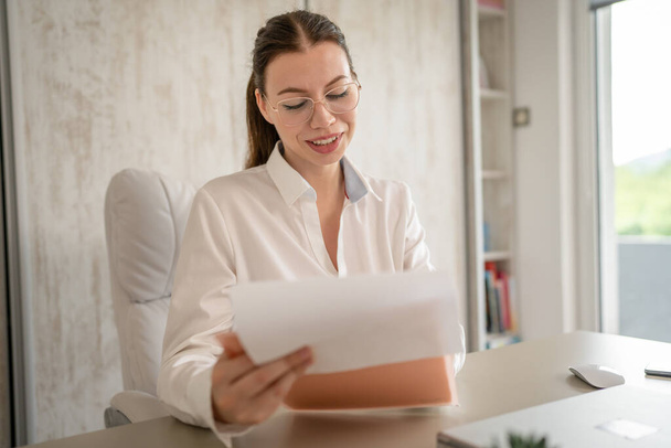 Beyaz gömlekli bir iş kadını ya da sekreteri masasında oturmuş mutlu bir şekilde gülümsüyor kağıt belgeleri kontratına bakıyor ve beyaz gömlek fotokopisi çekiyor. - Fotoğraf, Görsel