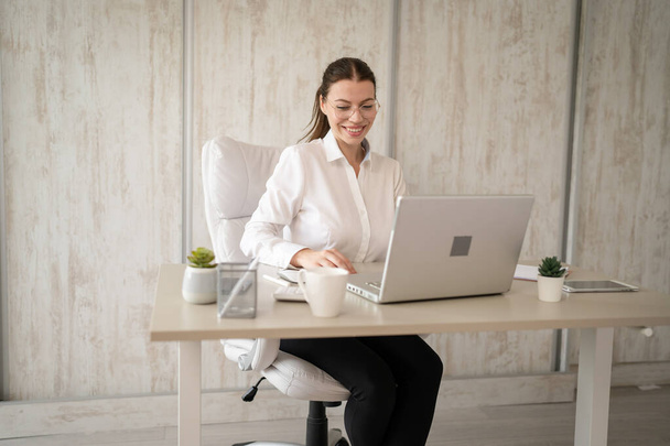 One woman female caucasian entrepreneur businesswoman or secretary sitting at her office at desk work on laptop computer happy smile wear white shirt copy space - Fotografie, Obrázek