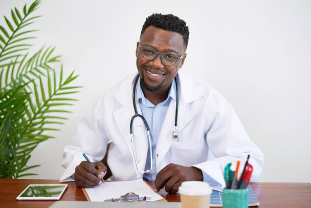 Ritratto di un medico del Black GP seduto alla scrivania a scrivere e sorridere. Foto di alta qualità - Foto, immagini