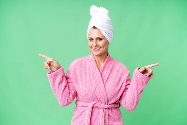 Middelbare leeftijd blanke vrouw in een badjas over geïsoleerde achtergrond wijzend vinger naar de lateralen en gelukkig - Foto, afbeelding