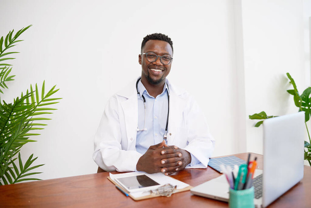 Portret czarnego lekarza rodzinnego siedzącego przy biurku z zapiętymi rękami. Wysokiej jakości zdjęcie - Zdjęcie, obraz
