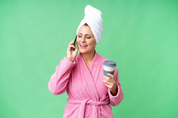 Middelbare leeftijd blanke vrouw in een badjas over geïsoleerde achtergrond houden van een gesprek met de mobiele telefoon - Foto, afbeelding