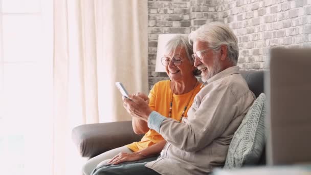 видео красивой романтической пожилой пары, использующей смартфон вместе на диване дома - Кадры, видео