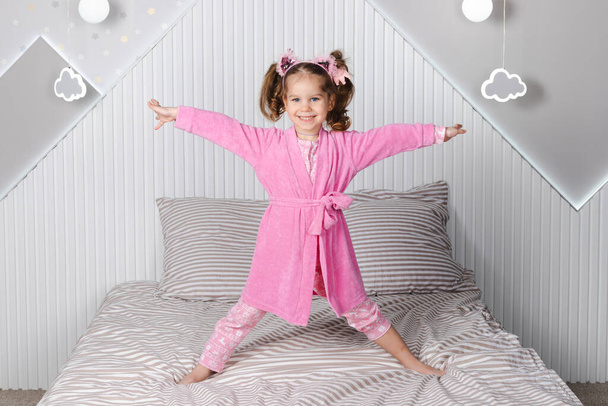 Ένα γοητευτικό κοριτσάκι με ροζ πιτζάμα και ρόμπα πηδάει στο κρεβάτι, με χαρά υψώνει τα χέρια της και γελάει.. - Φωτογραφία, εικόνα