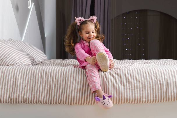 Ένα γοητευτικό κοριτσάκι με ροζ πιτζάμα και ρόμπα κάθεται στο κρεβάτι, φοράει τις μαλακές παντόφλες της και χαμογελάει.. - Φωτογραφία, εικόνα