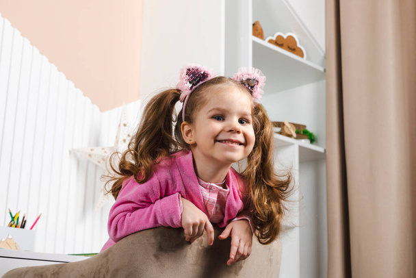 Lähikuva Viehättävä pieni tyttö vaaleanpunainen pyjama hymyilee ja katselee ulos reunasta nojatuoli. - Valokuva, kuva