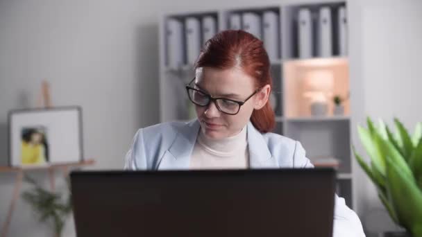 πορτρέτο της γοητευτικής γυναίκας που εργάζεται σε έναν υπολογιστή, ενώ κάθεται στο γραφείο στο γραφείο του σπιτιού της, χαμογελώντας και κοιτάζοντας κάμερα - Πλάνα, βίντεο
