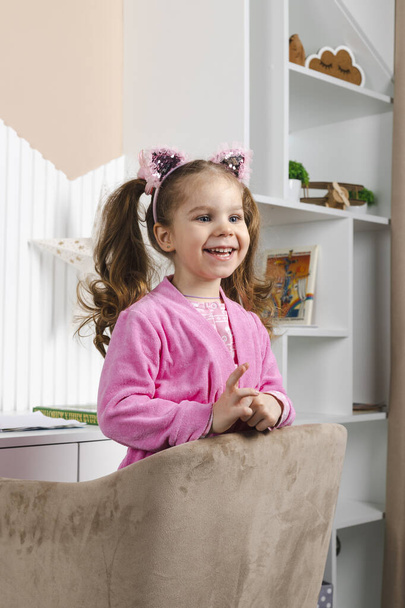 ピンクのパジャマ姿のチャーミングな女の子が笑顔でアームチェアの端から外を見ている. - 写真・画像