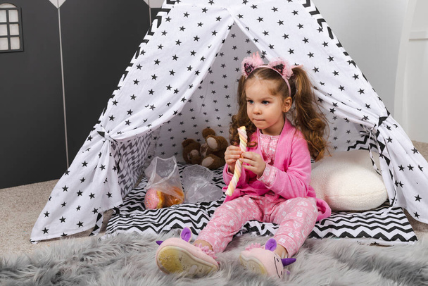 A charming little girl in a pink pajama and robe is sitting at home in a homemade childrens tent with toys and eating sweets and glazed buns. - Foto, Imagen