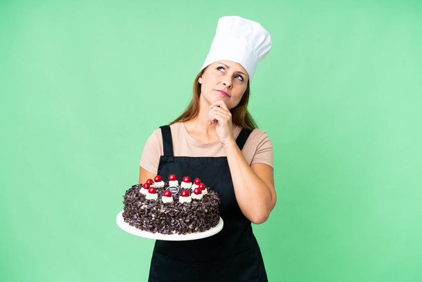 Кухар середнього віку жінка тримає великий торт над ізольованим тлом і дивиться вгору
 - Фото, зображення