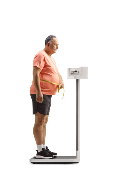 Full length profile shot of a corpulent mature man standing on a medical weight scale and measuring waste isolated on white background - 写真・画像