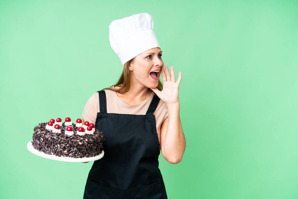 Μεσήλικας ζαχαροπλάστης γυναίκα κρατώντας ένα μεγάλο κέικ πάνω από απομονωμένη φόντο φωνάζοντας με το στόμα ορθάνοιχτο στο πλάι - Φωτογραφία, εικόνα