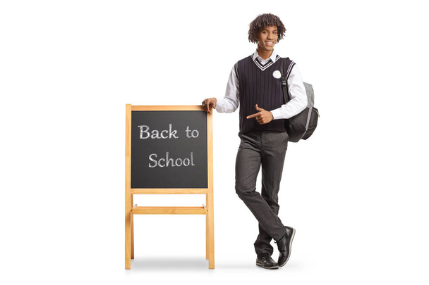 Full length portrait of a male african american student in a uniform leaning on blackboard with text back to school and pointing isolated on white background - Zdjęcie, obraz