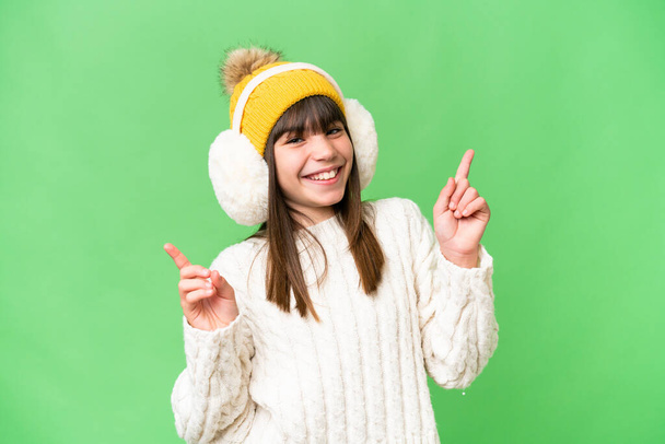 Pequeña chica caucásica con manguitos de invierno sobre fondo aislado señalando el dedo a los laterales y feliz - Foto, imagen