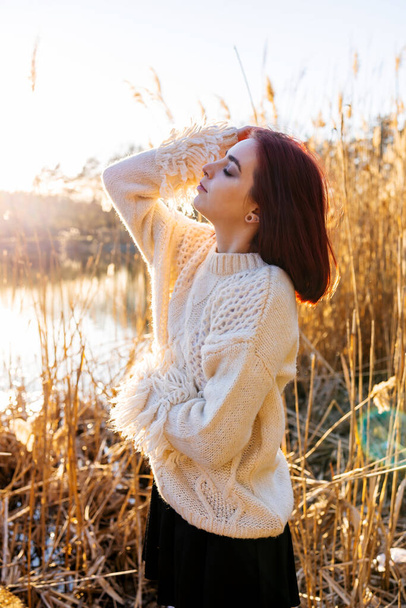 Elegant young woman in a white woolen sweater poses in the autumn park on the background of reeds. Autumn fashion and beauty. - Photo, Image