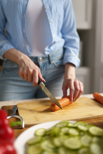 Woman cuts carrot for salad on a wooden cutting board. Female person cooking healthy vegetarian meal in a domestic kitchen - Photo, image