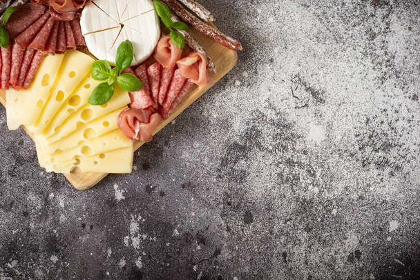 Delicacies. Cheese, prosciutto, salami on a wooden square board on a concrete background. - 写真・画像