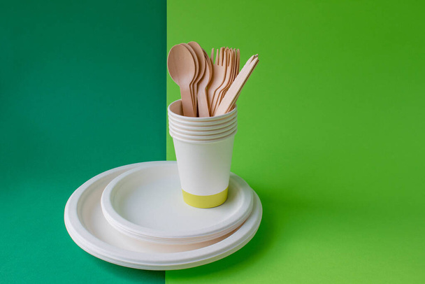Οικολογικά αναλώσιμα πιάτα από ξύλο μπαμπού και χαρτί σε πράσινο φόντο. Κουτάλια, πιρούνια, μαχαίρια, μπολ μπαμπού με χάρτινα φλιτζάνια - Φωτογραφία, εικόνα