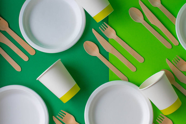 Eco-friendly disposable dishes made of bamboo wood and paper on a green background. Draped spoons, forks, knives, bamboo bowls with paper cups, abstract - Photo, Image