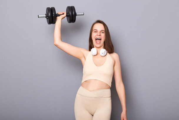 Portrait of crazy excited strong woman wearing top and leggins holding barbell in hand raised her arm doing exercise for her triceps and biceps isolated on gray background - Fotografie, Obrázek