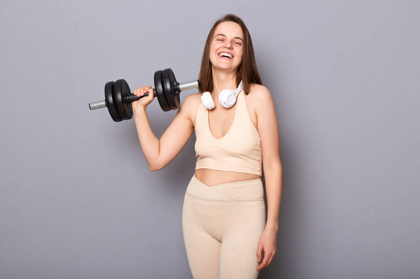 Indoor shot of laughing funny young woman wearing sportswear holding dumbbell making weightlifting training biceps and triceps with pleasure isolated on gray background - Fotó, kép