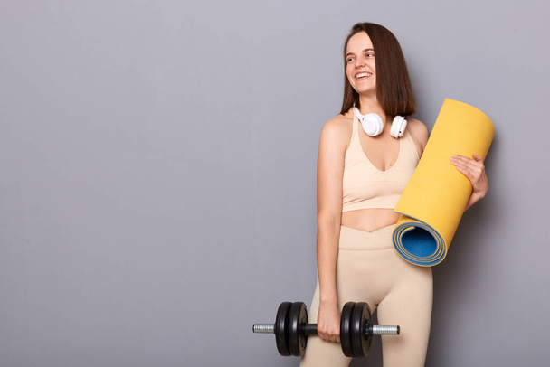 Portrait of smiling attractive young woman wearing sportswear holding dumbbell and rolled karemat looking away at advertisement area for gym isolated on gray background - Fotoğraf, Görsel