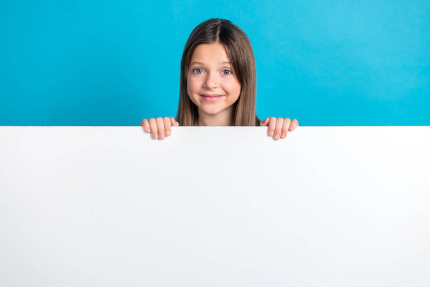 Photo of pleasant optimistic schoolgirl with straight hairdo dressed white shirt hold poster empty space isolated on blue color background. - Φωτογραφία, εικόνα
