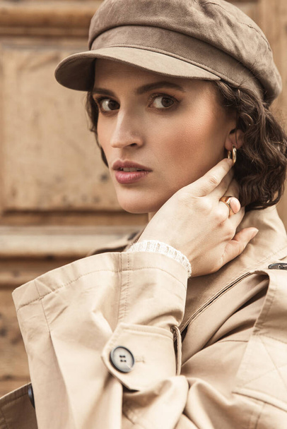 Young beautiful girl, brunette with curly hair. Dressed in a stylish beige trench coat and cap. - Photo, image