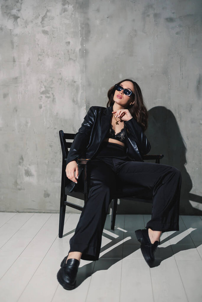 A cute young woman in a leather jacket and black pants wearing sunglasses is sitting on a chair on a gray background. - Foto, imagen
