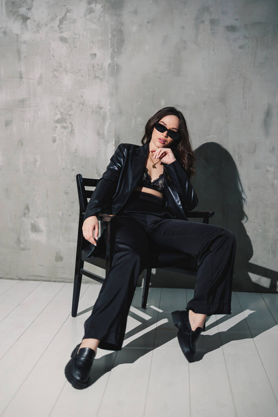A cute young woman in a leather jacket and black pants wearing sunglasses is sitting on a chair on a gray background. - Fotografie, Obrázek