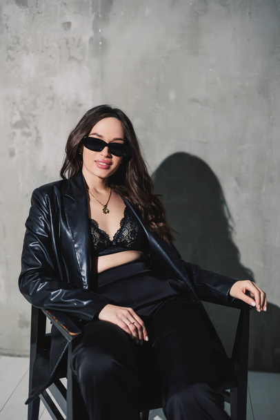 Eine niedliche junge Frau in Lederjacke und schwarzer Hose mit Sonnenbrille sitzt auf einem Stuhl vor grauem Hintergrund.. - Foto, Bild