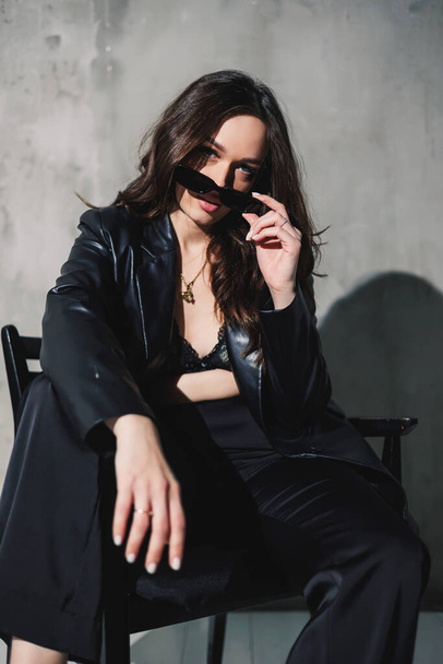 A cute young woman in a leather jacket and black pants wearing sunglasses is sitting on a chair on a gray background. - Foto, Bild