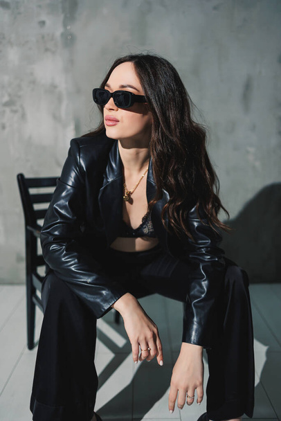 Uma jovem mulher bonita em uma jaqueta de couro e calças pretas usando óculos de sol está sentada em uma cadeira em um fundo cinza. - Foto, Imagem