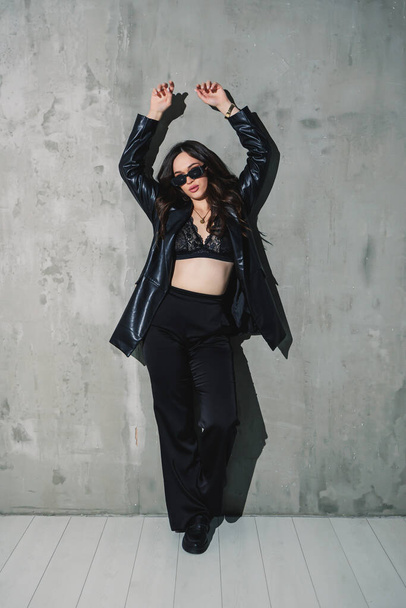 Beautiful elegant young woman model in nice leather suit, pants, jacket, stylish sunglasses posing on gray background. - Фото, изображение