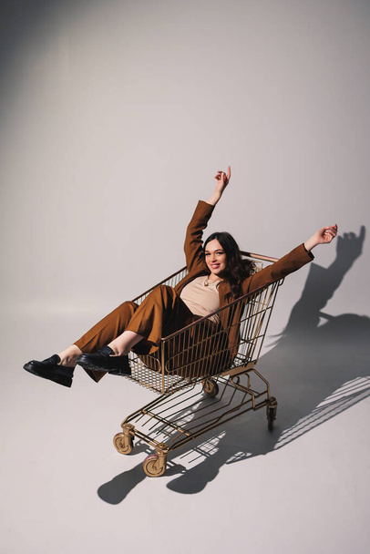 Beautiful elegant young woman model in nice stylish brown suit posing in shopping cart on gray background. Young elegant woman in a suit - Zdjęcie, obraz