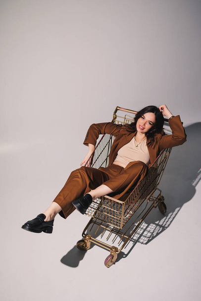 Beautiful elegant young woman model in nice stylish brown suit posing in shopping cart on gray background. Young elegant woman in a suit - Φωτογραφία, εικόνα