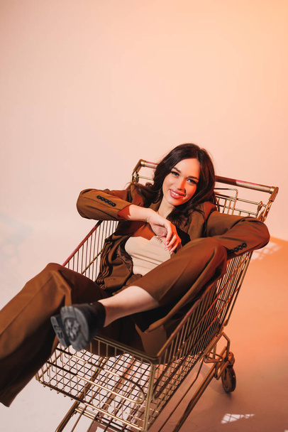 Beautiful elegant young woman model in nice stylish brown suit posing in shopping cart on gray background. Young elegant woman in a suit - Photo, Image