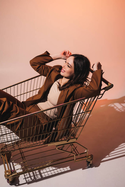 Schöne elegante junge Frau Modell in schönen stilvollen braunen Anzug posiert in Einkaufswagen auf grauem Hintergrund. Junge elegante Frau im Anzug - Foto, Bild