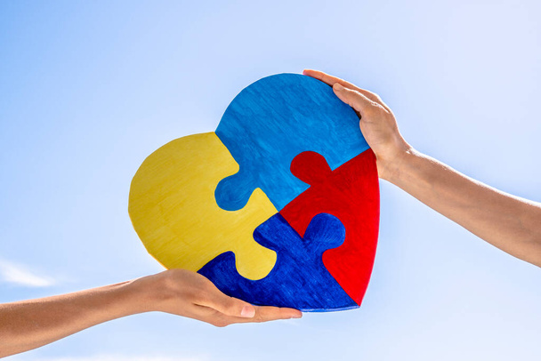 Světový den autismu, koncepce autismu. Dospělé a dětské ruce drží pohromadě barevné malované puzzle srdce na modrém pozadí oblohy. - Fotografie, Obrázek