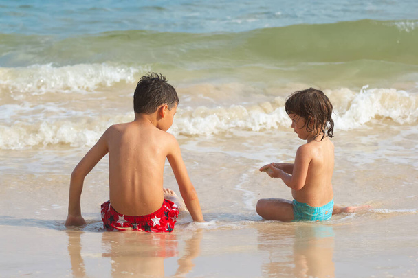 Tyły dwójki dzieci, brata i siostry, którzy siedzą na piasku nad brzegiem morza. - Zdjęcie, obraz