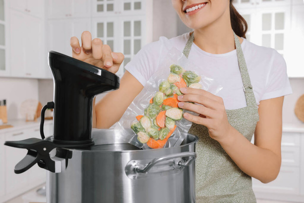 Woman putting vacuum packed vegetables into pot and using thermal immersion circulator in kitchen, closeup. Sous vide cooking - Photo, image